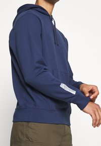 Navy blue hoodie made of soft material, featuring a front pocket, drawstring hood, and a label detail on the left sleeve.