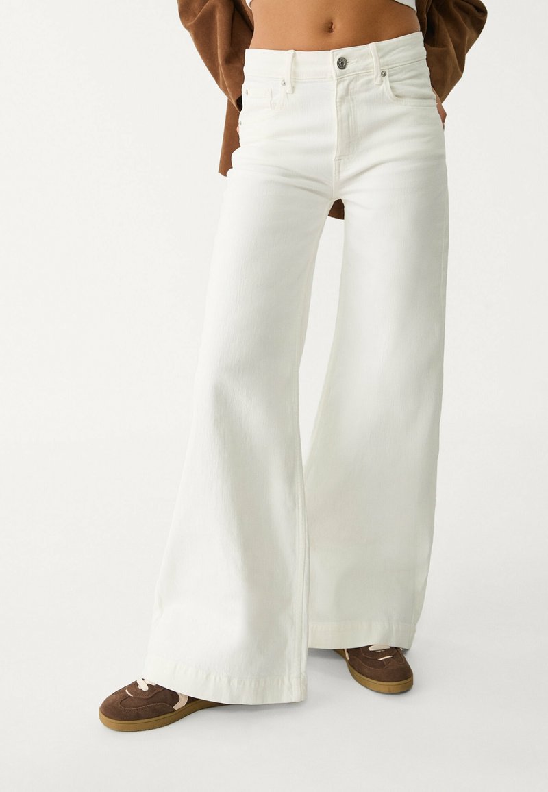 Femme portant un jean blanc taille haute, jambes larges, avec des baskets marron et une veste marron courte, sur un fond uni.