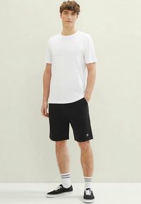 Weißes T-Shirt mit kurzen Ärmeln und Rundhalsausschnitt, kombiniert mit schwarzen Shorts mit Logo-Akzent. Schwarze Sneaker und weiße Sportsocken vervollständigen das Outfit.
