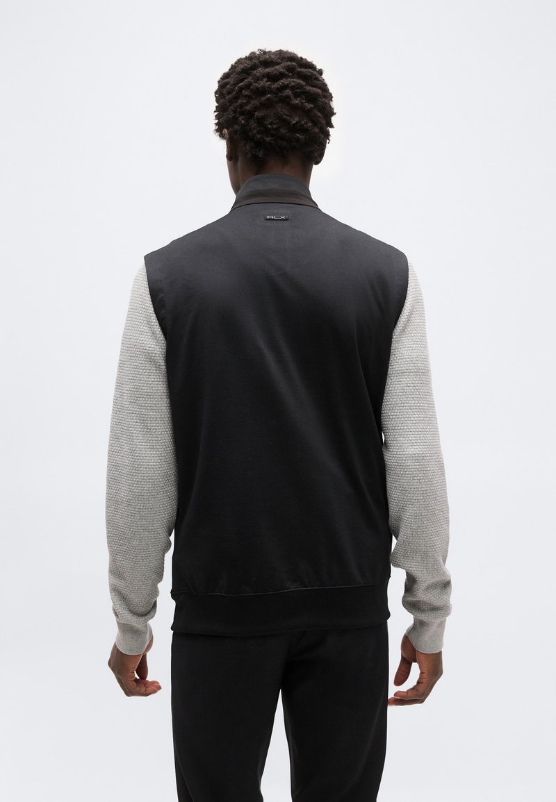 Homme aux cheveux courts et bouclés portant un gilet noir sans manches par-dessus une chemise gris clair à manches longues texturée, vu de dos sur un fond uni.