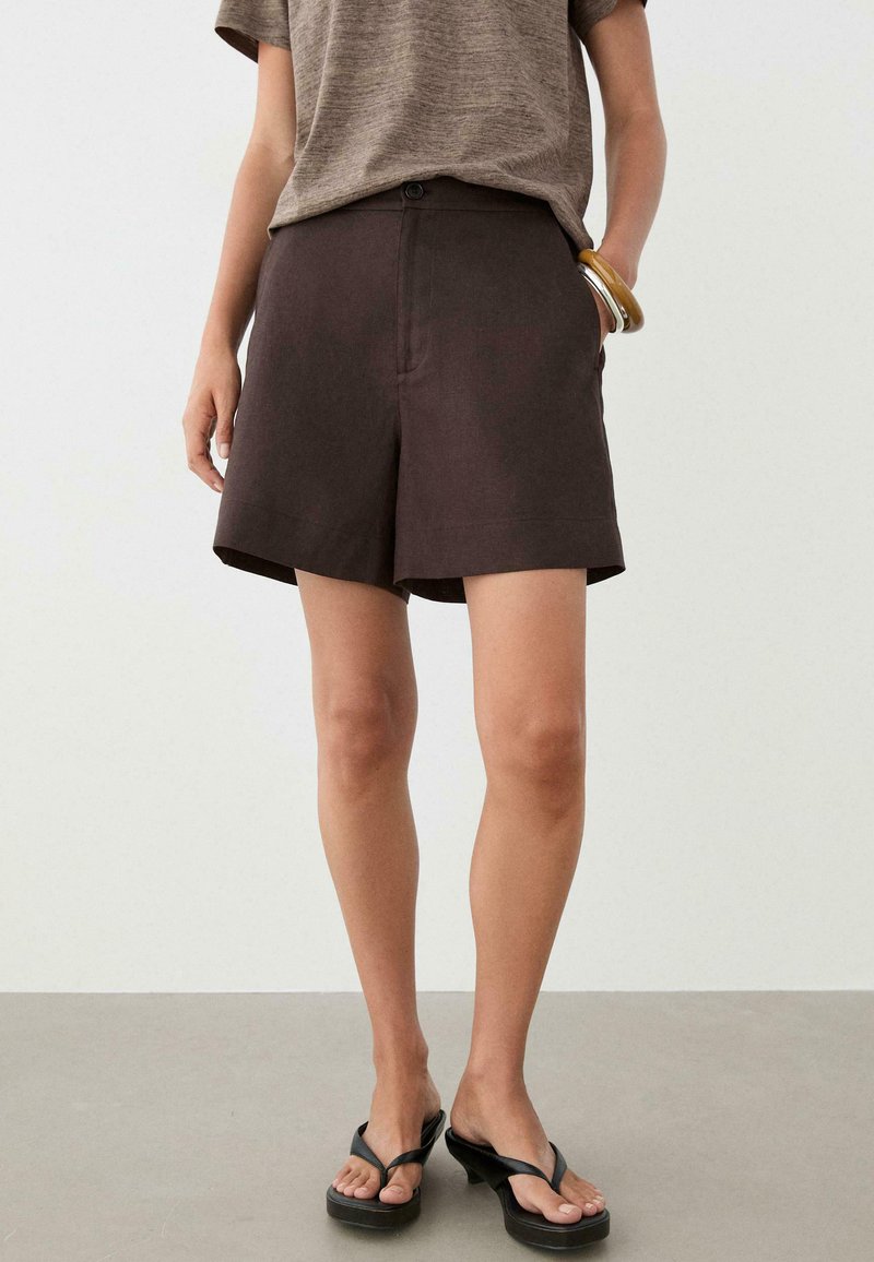 Person wearing dark brown tailored shorts, taupe short-sleeve shirt, black heeled sandals, and chunky bracelets on one wrist, standing on gray floor.
