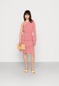 Vestido rosa assimétrico com design em camadas, feito de tecido leve. Acompanhado de uma mala amarela com padrão e sandálias amarelas de dedo aberto.