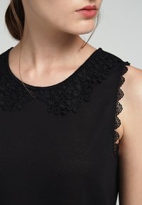 Černý top se strukturovaným krajkovým výstřihem s květinovými vzory. Látka je hladká a design má klasický, bezramenný střih.