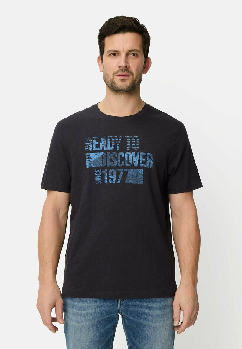 Sort kortærmet t-shirt lavet af bomuld, med et blåt grafisk tryk og tekst. Model iført blå jeans, neutral baggrund.