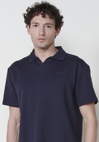 Polo à manches courtes bleu marine en tissu texturé, avec un col en V et un subtil logo brodé sur la poitrine gauche .