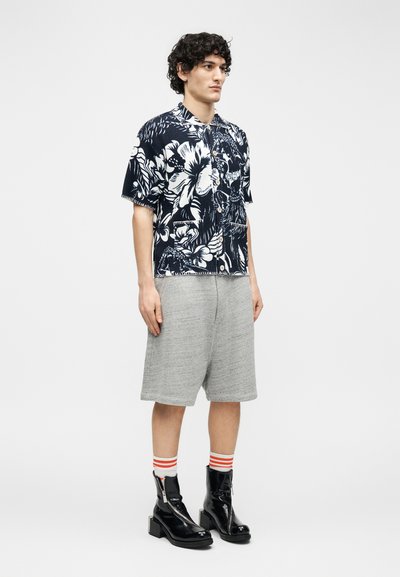 Mladi mož stoji in nosi črno-belo cvetlično srajco s kratkimi rokavi, sive kratke hlače do kolen, črtaste nogavice in črne gležnjarje.