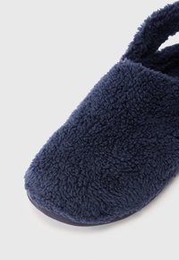 Nära bild av marinblå fuzzyslipper med plyschtextur och mjuk sula på en vit bakgrund.