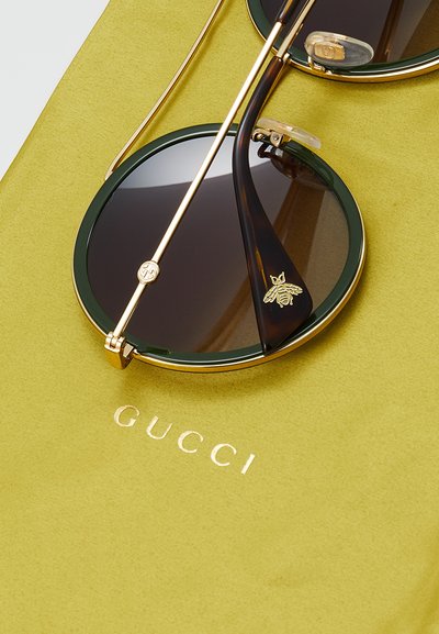 Ringikujulised Gucci prillid tumedate läätsede, rohelise raamiga, kuldsete detailide ja mesilase motiiviga. Asetsetud pehmele kollasele kangale, millel on embosseeritud logo.