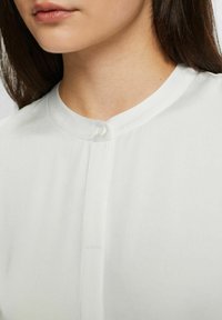 Bílá košile s knoflíky a kulatým výstřihem, s malým zapínáním na knoflík a hladkou, lehkou texturou látky.