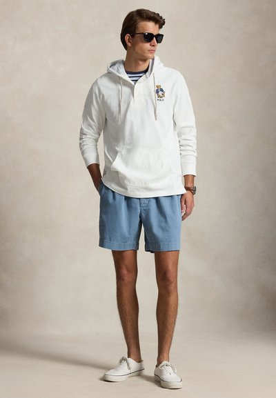 Homme portant un sweat à capuche blanc avec un logo d'ours, un short bleu, des baskets blanches, des lunettes de soleil et une montre, debout les mains dans les poches sur un fond uni.