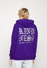 Sudadera morada con un gran gráfico plateado que dice "KINDNESS" en la parte posterior. Presenta un bolsillo frontal tipo canguro y puños de canalé.