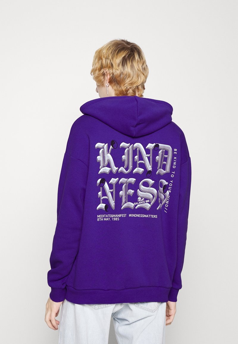 Sudadera morada con un gran gráfico plateado que dice "KINDNESS" en la parte posterior. Presenta un bolsillo frontal tipo canguro y puños de canalé.