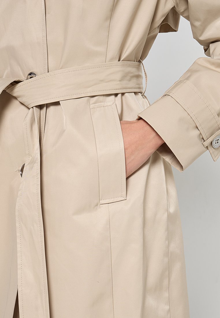 Trench-coat beige, imperméable, avec une taille ceinturée, des épaulettes, des poches latérales et des poignets boutonnés, présentant une texture lisse et brillante.