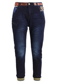 Donkere denim jeans met geborduurde details op de zakken. Voorzien van een patroon op de tailleband en gestreepte boorden in geel en zwart.