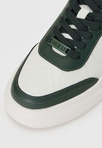 White sneaker with green accents, textured leather upper, prominent logo on the tongue, and a thick rubber sole. Distinct stitching detail.