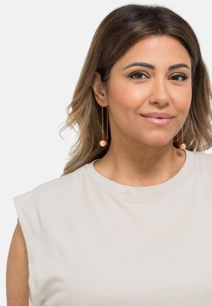 Roségoldene Tropfohrringe mit sphärischen Akzenten, die eine zarte Kette aufweisen. Das Model trägt ein schlichtes ärmelloses Shirt in Hellgrau.