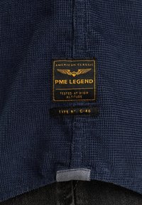 Tamno plavi teksturirani materijal s istaknutom oznakom brenda koja glasi "PME LEGEND." Oznaka sadrži zlatne akcentе i detalje o vrsti proizvoda.