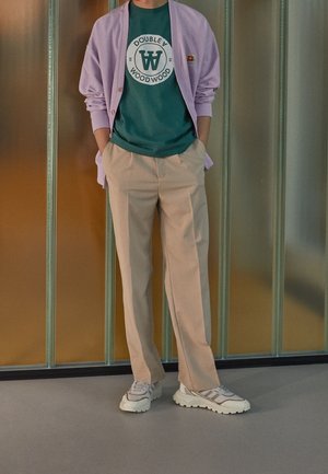 Osoba stoi z rękami w kieszeniach, ubrana w fioletowy sweter rozpinany, zielony T-shirt z nadrukiem, beżowe spodnie i białe masywne sneakersy.