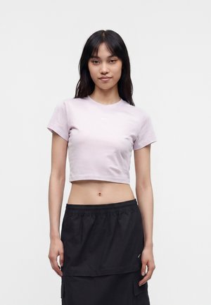 Jeune femme aux longs cheveux foncés portant un t-shirt court rose clair et une jupe noire, debout devant un fond blanc uni.