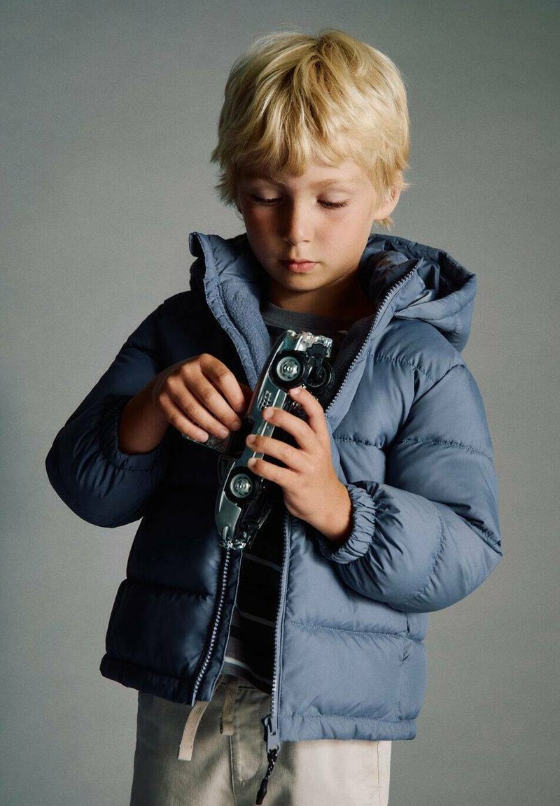 Doudoune bleue rembourrée avec capuche ; un enfant tient une voiture en jouet argentée, portant un t-shirt gris à rayures horizontales et un pantalon clair.