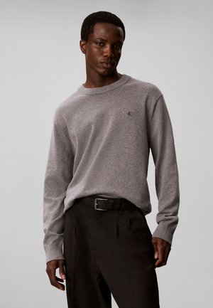 Homme portant un pull gris Calvin Klein et un pantalon noir, debout devant un fond uni gris clair.