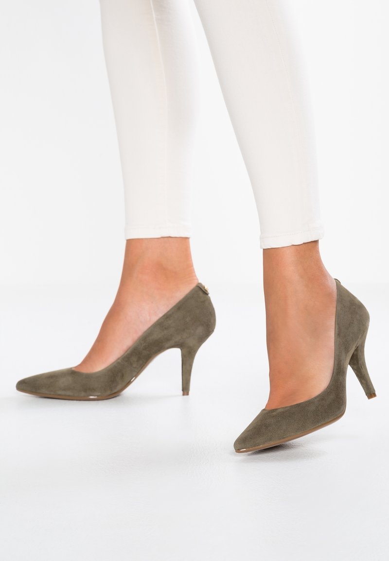 Femme portant un pantalon beige slim et des talons hauts pointus en daim vert olive, debout sur un sol blanc.