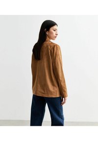 Bruine suède jas met een relaxte pasvorm, lange mouwen en minimale stikdetails, gecombineerd met donkerblauwe wijduitlopende jeans.