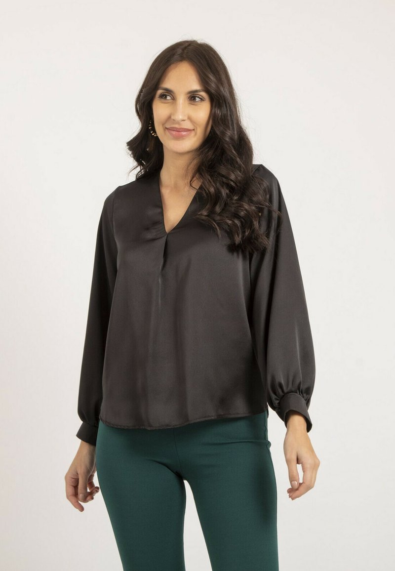 Blouse en satin noir avec un col en V, des manches longues ballon et une coupe ample. Portée avec un pantalon vert ajusté, sur un fond clair.
