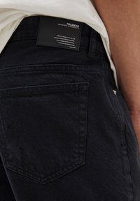 Jeans de mezclilla negra con un tejido texturizado, que cuentan con un bolsillo trasero y una etiqueta de parche de cuero negro en la cinturilla con texto blanco.