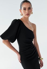 Sort asymmetrisk kjole med tekstureret stof, voluminøs pufærme, rynket detalje og en figursyet silhuet. Guld manchetarmbånd som accent.