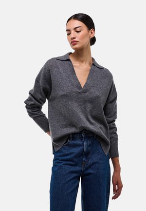 Pull en V gris avec un design à col, coupe légèrement oversize, poignets côtelés et fentes latérales, assorti à un jean bleu taille haute.