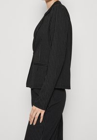 Donna vestita con un completo a righe nere, con maniche lunghe, tasche laterali e pantaloni coordinati, in piedi di fronte a uno sfondo bianco uniforme.