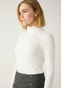 Langärmliges, weißes Rollkragenoberteil aus glattem Stoff mit taillierter Silhouette. Das Model kombinierte es mit einem grauen, hoch taillierten Rock.