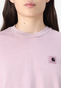 Lyserød bomulds t-shirt med ribbet crew neck og et kvadratiske logo patch på venstre bryst med et sort design. Glat tekstur.