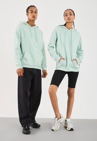 Piparmētru zaļa kapuce ar kangarū mīkstajām kabatām un auklām, kombinēta ar melniem džinsiem un velobiksēm. Ikdienišķi sportiski apavi noslēdz izskatu.