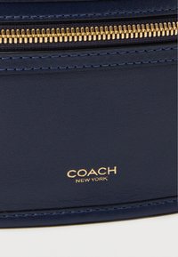 Temnomodra usnjena torbica z zlatim zadrgo. Na njej je vtisnjen logotip "COACH NEW YORK" v zlatih črkah. Ima teksturirano površino in kontrastno šivanje.