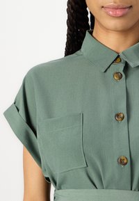 Abito camicia verde con colletto, maniche corte a risvolto e chiusura a bottoni, dotato di taschino sul petto e texture liscia.