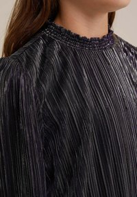 Svart plisserad topp med en hög, volangkrage. Tyget har subtila metalliska ränder i lila och silver, vilket skapar en texturerad look.