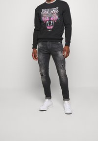 Černá mikina s barevným motivem tygra a kulatým výstřihem, v kombinaci s roztrhanými šedými úzkými džíny a bílými teniskami s perforacemi.