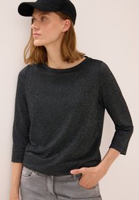 Schwarzes, strukturiertes Langarmshirt mit Rundhalsausschnitt und Dreiviertelärmeln, kombiniert mit grauen Hosen. Es wird eine schwarze Mütze getragen.