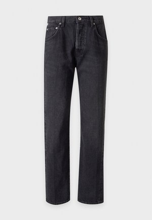 Femme portant un col roulé sans manches rayé, un pull noir posé sur les épaules, un jean noir et des mocassins noirs, debout devant un fond uni.