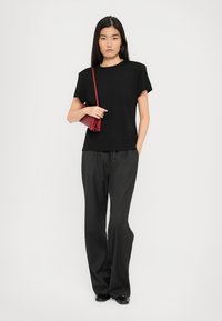 Woman standing wearing a black t-shirt, loose black pants, black shoes, and holding a small red shoulder bag.