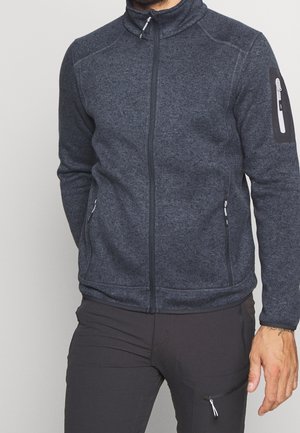 Mężczyzna ubrany w ciemnoszarą polarową kurtkę na zamek z zasuwanymi kieszeniami z przodu i na rękawie, w połączeniu z czarnymi spodniami z kieszenią na udzie zapinaną na zamek.