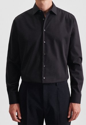 Mann trägt ein eng anliegendes, schwarzes, langärmliges Hemd mit Knöpfen, in schwarze Hose gesteckt, steht vor einem einfarbigen Hintergrund.