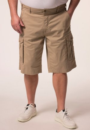 Mann trägt beige Cargo-Shorts und weiße Sneaker, steht mit teilweise in den Taschen befindlichen Händen vor einfarbigem Hintergrund.