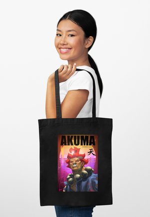 Młoda kobieta uśmiecha się, trzymając czarną torbę typu tote z kolorową grafiką "AKUMA Street Fighter" z dziką rudowłosą postacią.