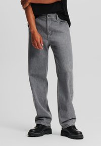 Szürke denim farmer, laza fit, öt zsebes kivitel, cipzáras záródással. Fekete fűzős csizmával viselve. Sima textúra, laza stílus.