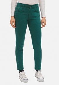 Tejanos elásticos verdes de algodón con un corte slim, que cuentan con cinco bolsillos, un cierre de botón y longitud hasta el tobillo, combinados con zapatillas blancas.