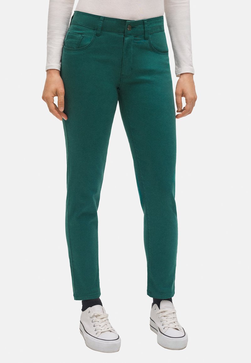 Tejanos elásticos verdes de algodón con un corte slim, que cuentan con cinco bolsillos, un cierre de botón y longitud hasta el tobillo, combinados con zapatillas blancas.