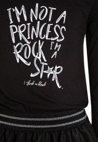 Černé tričko s dlouhým rukávem obsahující bílý textový grafický prvek a třpytivé akcenty. Elastický pas s kovovými pruhy na černé sukni.